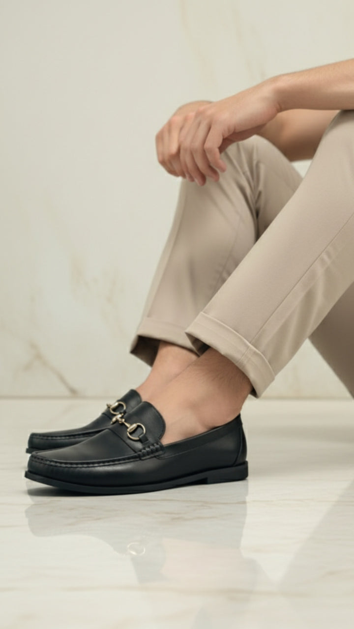 Person wearing black loafers with gold buckles on a light background